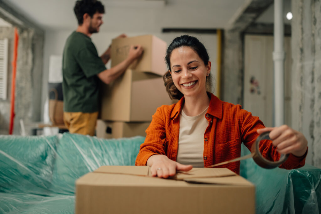 Smiling woman sealing a cardboard box while moving into a new home with her partner helping in the background